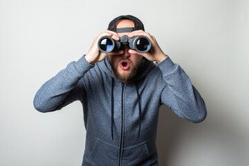 Young man with a surprised face in a hoodie and a cap looks through binoculars on a light background