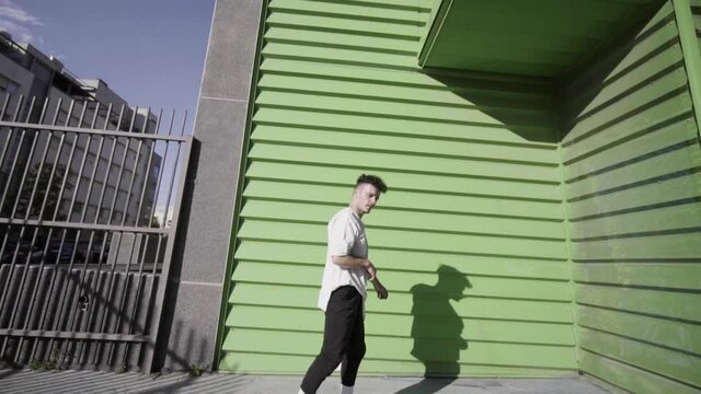 frontal shot traveling movement Caucasian boy curly black hair with short white t-shirt and long black sports pants with gray sports shoes doing parkour and flips free running in building green colors