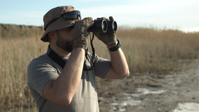 Side view of bearded adult man, hunter of a guard in military t-shirt and hat, looking through binoculars, secure territory, hunting in field, searching for something