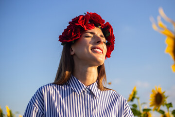 Young woman with closed eyes wearing a wreath of red flowers on a sunflower field at sunset