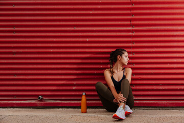 Runner rests leaning against a red wall. Athlete girl with sportswear next to her water bottle on the street