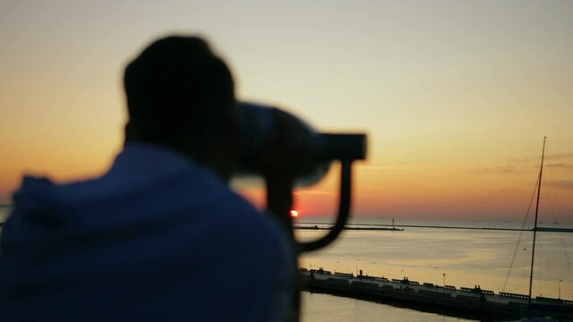 blurry man silhouette looks at orange seascape reflecting summer sunset through binoculars slow motion backside view
