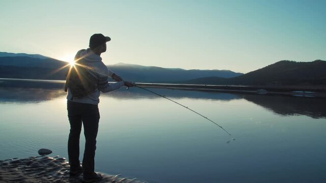 Medium silhouette shot of a fisherman casting his lure into a still, glassy, lake in front of gorgeous, snow-capped mountains.  The sun flares and highlights his cast.