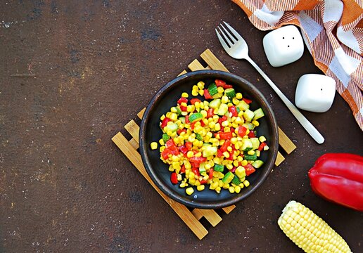 Vegetable stew or warm salad made from sweet corn, zucchini and red bell pepper on a clay dish on a brown concrete background. Vegan recipes.