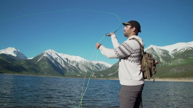 Medium-wide shot of a fly fisherman casting his lure into a deep blue lake in front of gorgeous, snow-capped mountains.  The fisherman casts several times before finally letting his lure land.