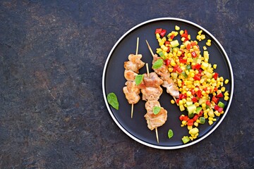 Chicken kebab on wooden skewers on a black plate on a black concrete background. Served with a warm salad of sweet corn, zucchini and red pepper. Thanksgiving Day.