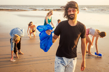 handsome indian man in the foreground collecting plastic from sand into the bag , his mixed race...