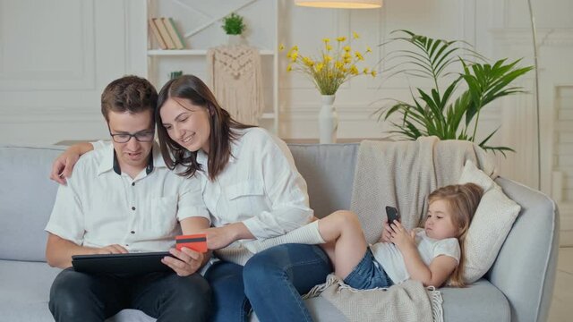 Parents Shopping Online While Their Daughter Plays in a Phone Online Game During a Sitting on the Couch at Home. Purchase Confirmation by the Internet. Binding a Card for Online Shopping