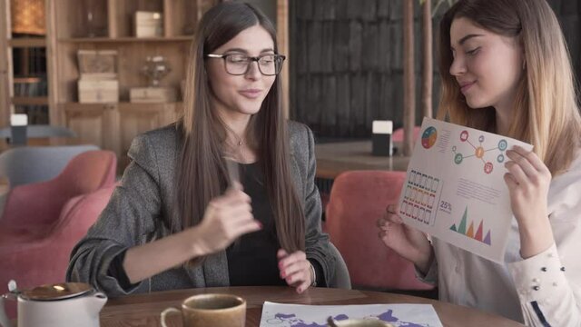 Two charming young woman are presentating some data and looking directly at the camera. One woman is holding a diagrams and other is explaining them.