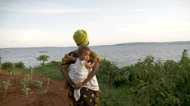 A Tracking Shot From Behind Of An African Woman In Traditional Cultural Clothes Walking With Her Baby On Her Back Through A Rural Village.