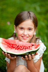 8-year-old girl in white clothes and with two tails on her head eats watermelon on the grass in the park