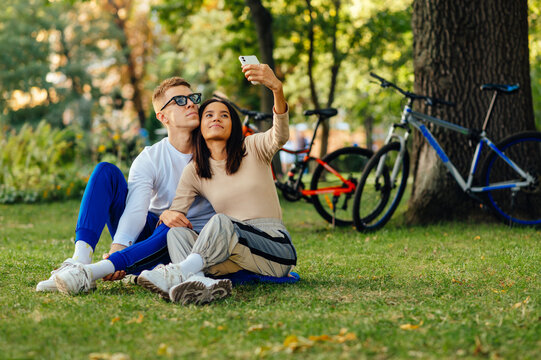 Beautiful young couple taking a selfie sitting in the park on the grass on a background of bicycles.Woman and man spend time together in the park, take a joint photo without a smartphone selfie camera