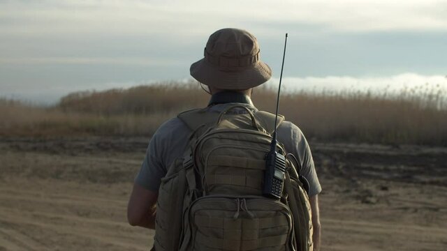 Slow motion rear view of bearded soldier, guard on duty with backpack and walkie-talkie, patrolling field, moving forward in plain area, checking territory for intruders