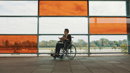Young independed caucasian woman in the wheeldchair reading a book in front of big glass...