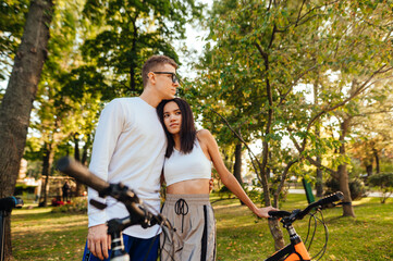 Beautiful stylish couple standing with bicycles in the autumn park and hugging, looking away. Man and woman in love with bicycles embracing on a walk.