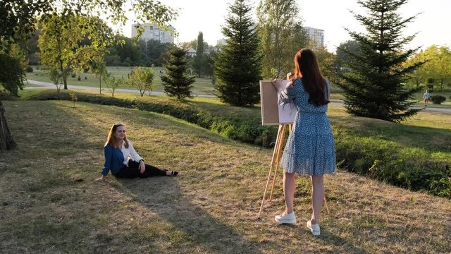 Pretty woman with red hair draws a portrait of a girl that is sitting on grass, in pencil, outside in the park.