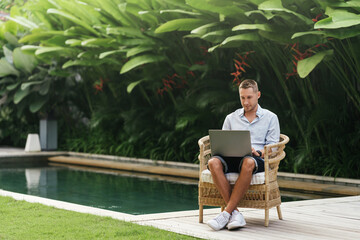 Young man using a laptop computer in a garden with a swimming pool. business, study, freelance