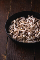 Tasty chopped champignon mushrooms in frying pan, uncooked food sliced, dark brown wooden background, angle view