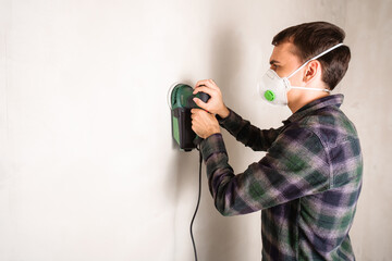 Man in protective mask working with electric sander to smooth plaster wall surface, room renovation...