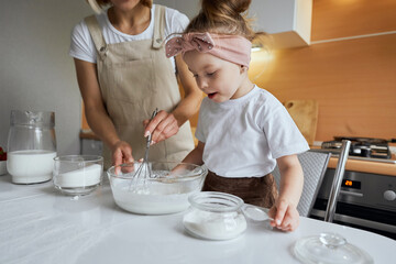 happy mother baking with little daughter in apron and cook hat working with flour , bowl and spoon preparing dough teaching the kid baking and having fun together.