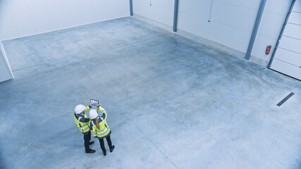 Industry 4.0 Modern Factory: Two Engineers in Empty Warehouse Use Digital Tablet Computer with Augmented Reality Software For a Room Mapping. Doing 3D Factory Floor Layout. High Angle Shot