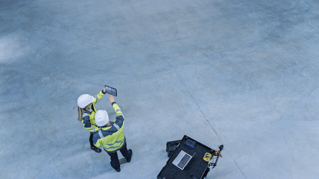 Industry 4.0 Modern Factory: Two Engineers Use Digital Tablet Computer With Augmented Reality Software Visualizing Of Manufacturing Plant Room Mapping. Doing 3D Factory Floor Layout. High Angle Shot