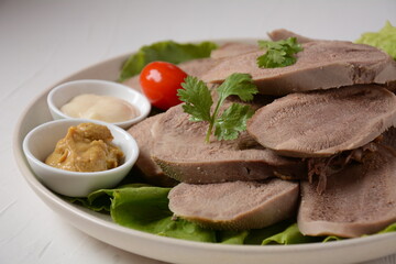 Sliced Beef Tongue Slices on a platter with lettuce leaves, cherry tomatoes and Dijon mustard on a wooden background