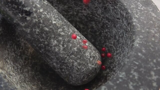 Rose-red peppercorns are falling into the grey stone mortar in slow motion