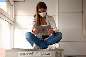 young woman wearing a protective mask sitting on a big washing machine and using a tablet.