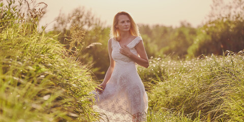 A blonde girl in a white dress walks outdoors in the high field grass