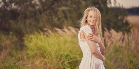 A blonde girl in a white dress walks outdoors in the high field grass