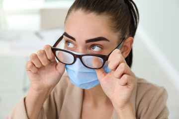 Woman wiping foggy glasses caused by wearing medical mask indoors, closeup