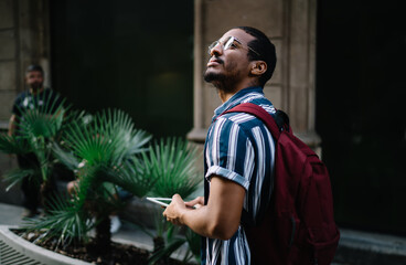 Positive dark skinned male traveler using digital tablet for searching location via navigation app walking on street, smiling african american with touchpad and rucksack visiting town during vacations