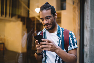 Positive dark skinned dark skinned hipster guy share content in blog satisfied with connection on street, smiling african american male influencer downloading content on smartphone outdoors