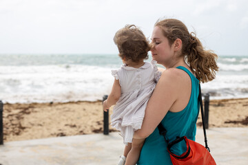 Fototapeta na wymiar a young mother gently lovingly holds a baby girl in her arms against the background of the sea