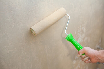 A man's hand with a paint roller is painting a wall with decorative plaster in beige. The concept of renovation of apartments and premises.