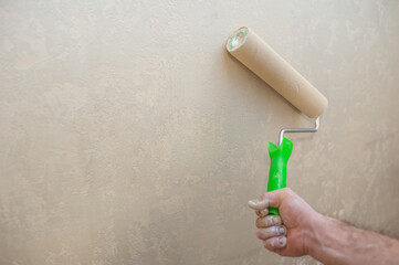 A man's hand with a paint roller is painting a wall with decorative plaster in beige. The concept of renovation of apartments and premises.