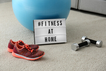 Sport equipment and lightbox with hashtag FITNESS AT HOME on floor indoors. Message to promote...