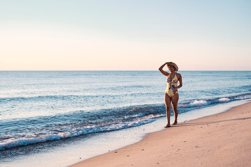 Latin girl walking near of the shore of a deserted beach. Wearing a yellow swimsuit and a hat. She is touching her hat.