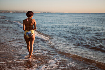 Latin girl walking near of the shore of a deserted beach. Wearing a yellow swimsuit. She is giving her back to the camera and watching the sunrise.