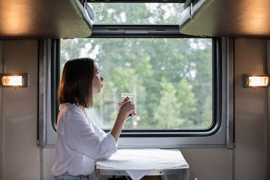 A young beautiful woman in a white shirt is sitting in a train compartment with a Cup of tea and looking out the window. Travel by train.