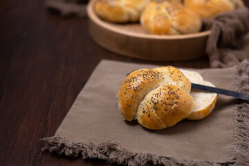 Close Up of a Homemade Kaiser Roll Topped with Poppy Seeds  on a Brown Napkin; More in a Round Wooden Bowl in the Background on a Wooden Table; Knife Used to Cut Roll Open