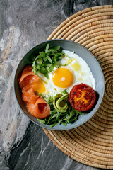 Healthy breakfast bowl with fried eggs, salmon, avocado, grilled tomato and salad serving with bread on straw napkin over dark marble background. Flat lay, copy space