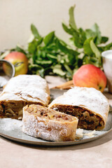 Homemade sliced classic apple strudel with icing sugar on ceramic plate with fresh apples, green leaves and cinnamon sticks above. Close up