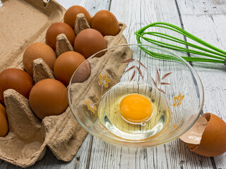 On a white board table stands an open package containing nine brown eggs. One egg is broken into a...