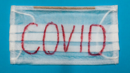 Typical white 3-ply surgical mask with rubber earhooks to cover mouth and nose with English block letters Covid on a blue background. Dangerous virus and quarantine concept.