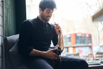Pensive bearded man carrying photo camera while sitting on windowsill in cafe