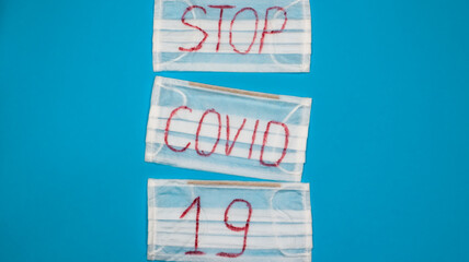 Typical 3-ply white surgical mask with rubber earhooks to cover mouth and nose with English block letters Stop Covid-19 on a blue background. Dangerous virus and quarantine concept. Copy space