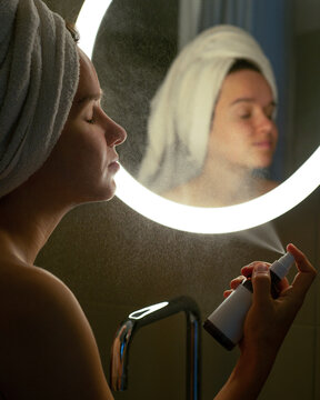 Young woman applying thermal water on face near mirror in bathroom.