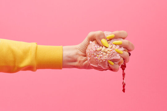 Woman with yellow nails squeezes a jammy iced pink doughnut in her hand